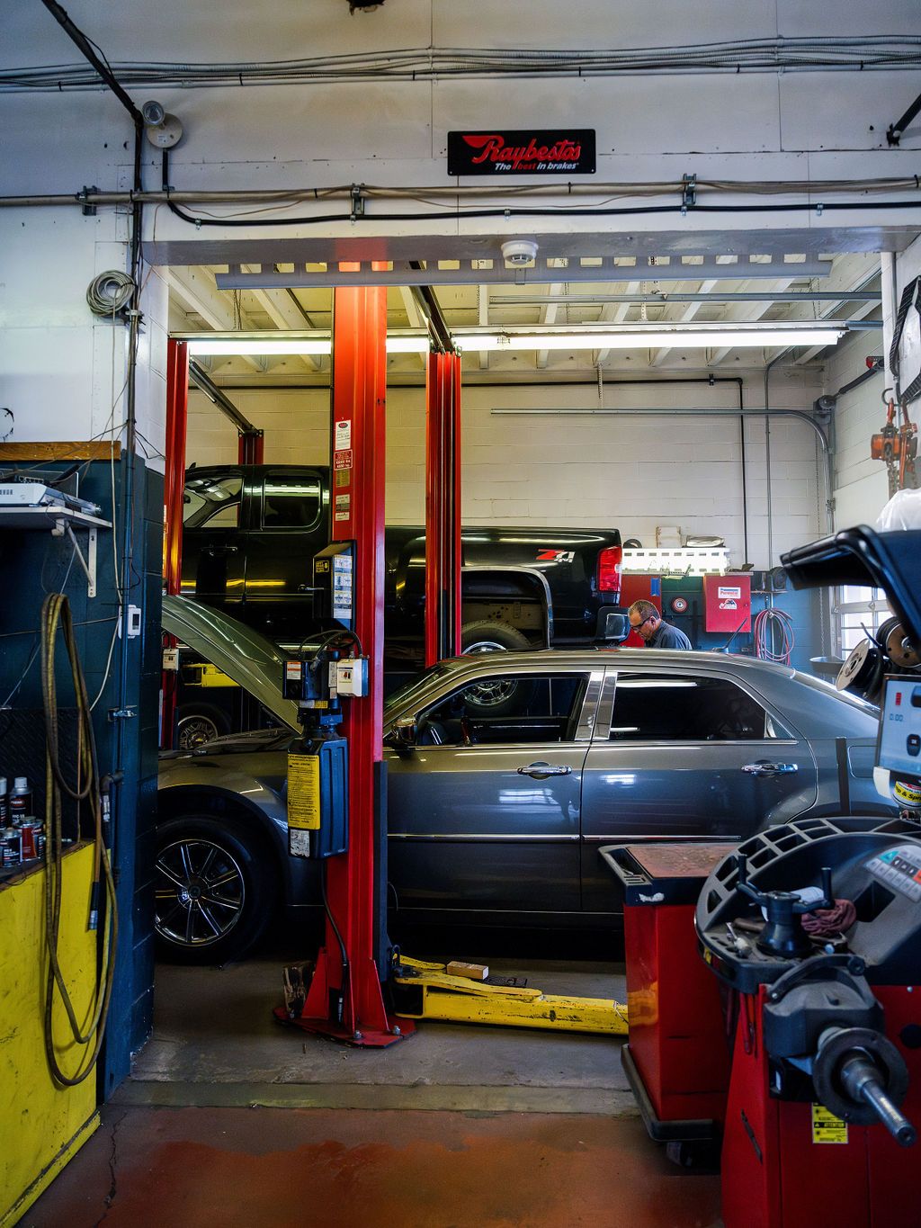 A grey car on a hoist in one of the shop's service bays, with a pickup truck raised on the hoist behind it