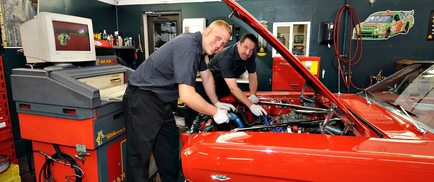 Two technicians working under the open hood of a red car beside diagnostic equipment in the service bay